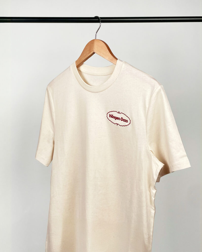 T-shirt beige personnalisé Häagen-Dazs avec logo brodé poitrine, textile promotionnel premium suspendu sur cintre.